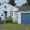 Kim's Cape prior to renovation shows blue vinyl siding, recessed entry slightly off-center and wrap-around corner windows.  Garage is the most prominent feature. 